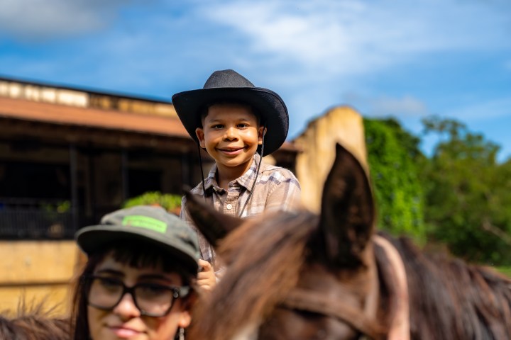 Smiling child in cowboy hat on horse, with woman and horse in foreground.