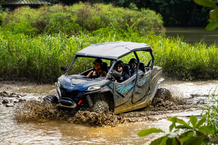 Four people in a side-by-side ATV drive through muddy water in a lush, green landscape.