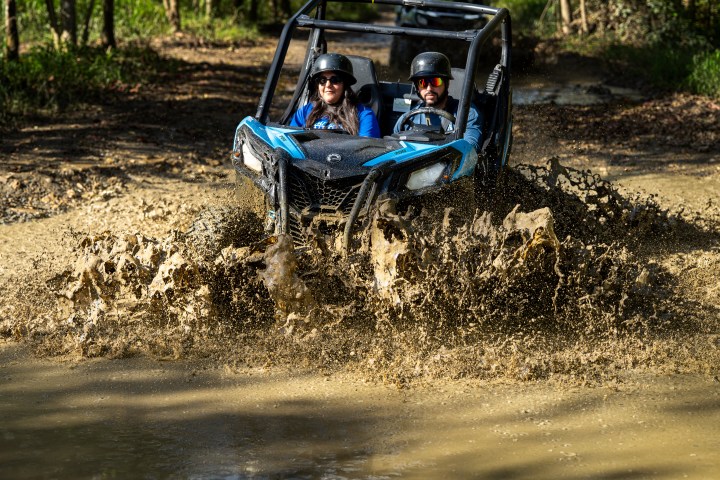 Two people in off-road buggy driving through muddy water in a forest.