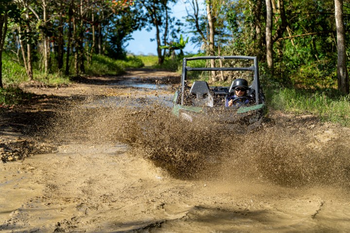 Person driving an off-road buggy through a muddy puddle in a forested area.