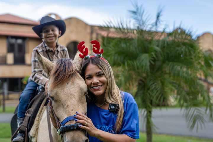 Child in cowboy hat on horse, woman with reindeer antlers beside them, in a garden setting.
