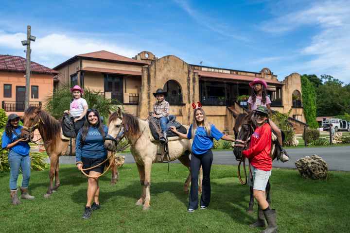 People and children on horses in front of a large building on a sunny day.